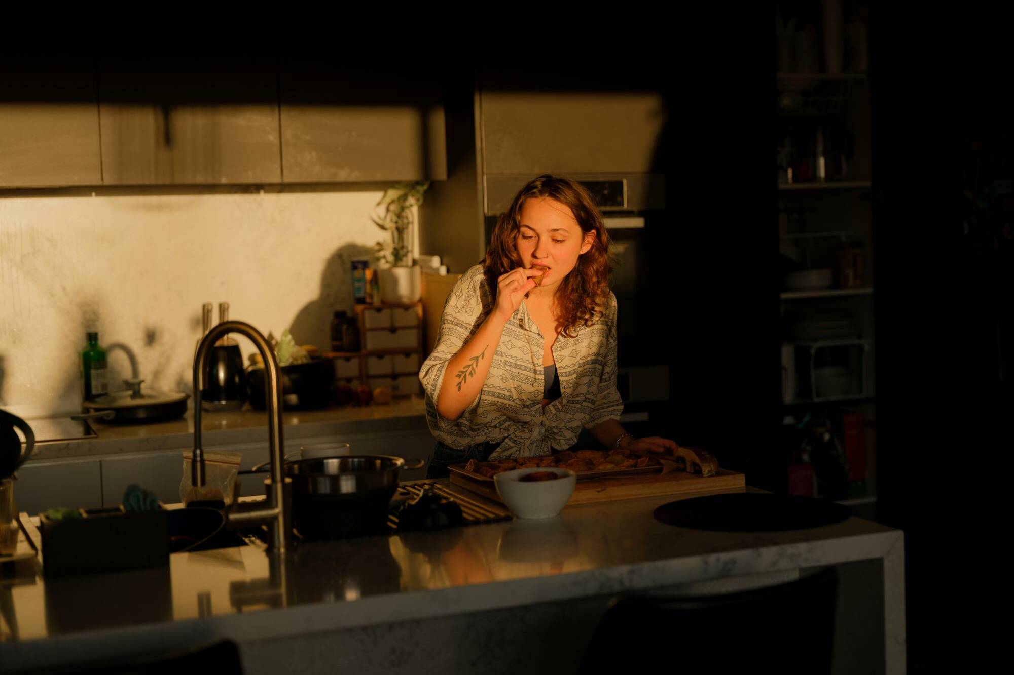 Femme debout dans une cuisine en fin de journée, mangeant avec les doigts pendant la préparation du repas