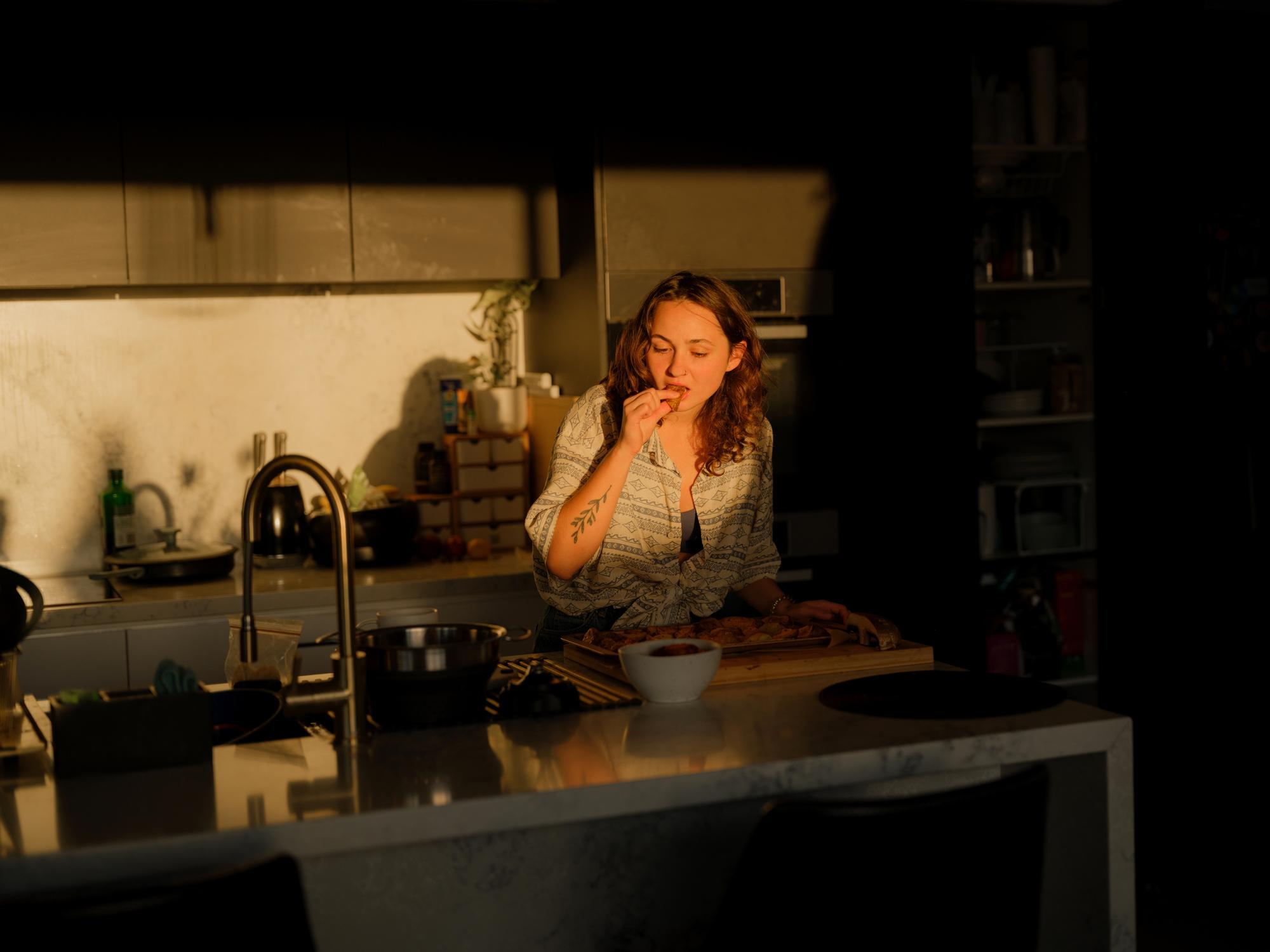 Femme debout dans une cuisine en fin de journée, mangeant avec les doigts pendant la préparation du repas